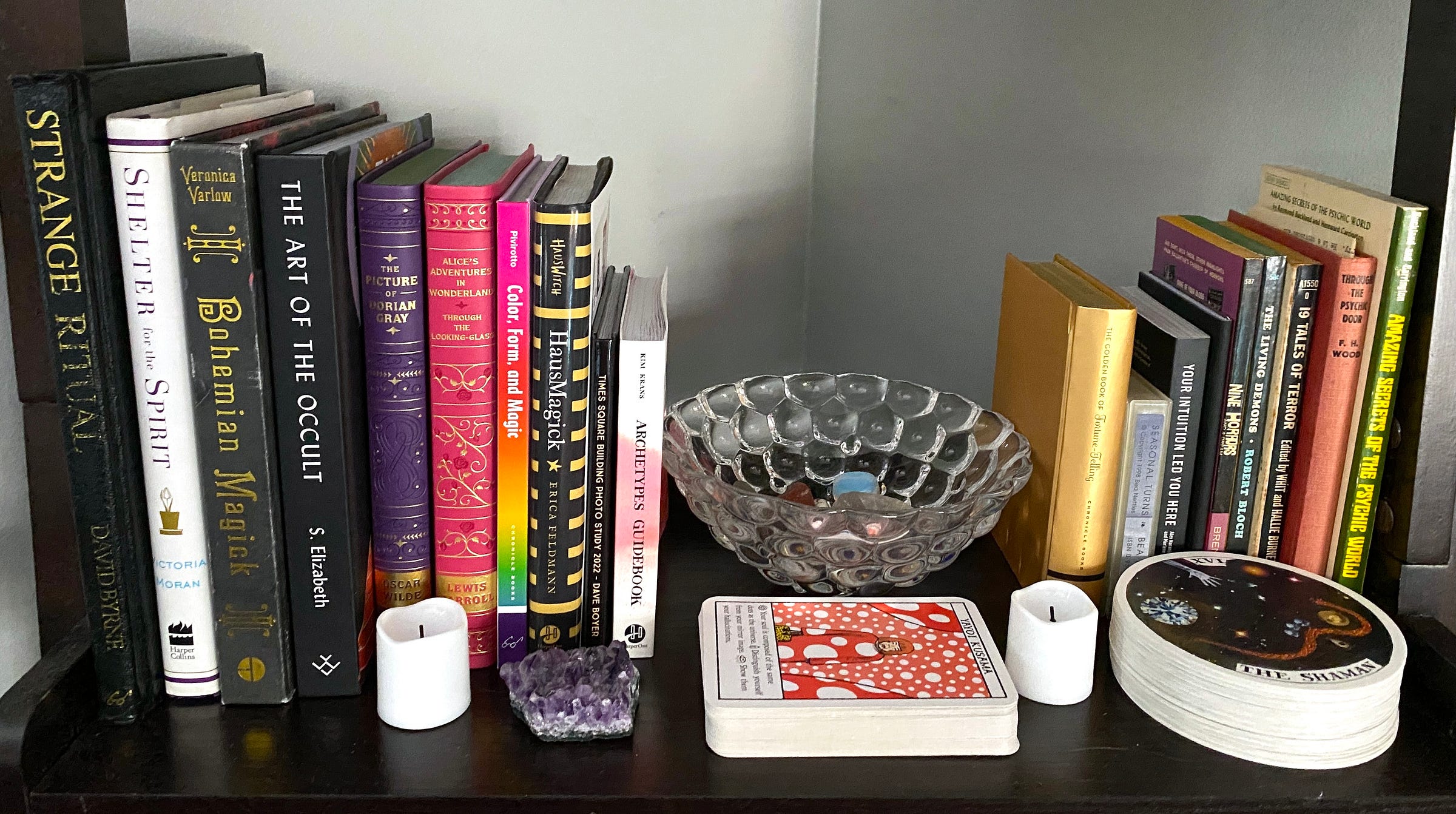 A shelf on a bookshelf in the author’s home, filled with books on magic and art, a glass dish filled with crystals, two plastic battery-powered votive candles, an amethyst crystal, and two oracle decks: the Wild Unknown Archetypes deck (card facing up: The Shaman) and the Artist Oracle (card facing up: Yayoi Kusama).