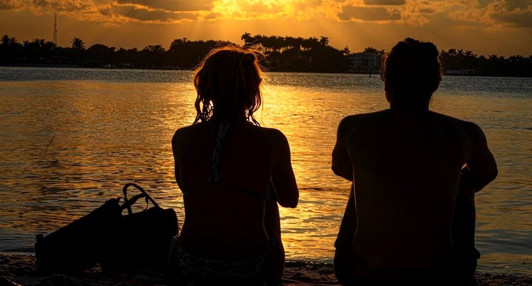 two people sitting on a beach watching the sun set two people sitting on a beach watching the sun set