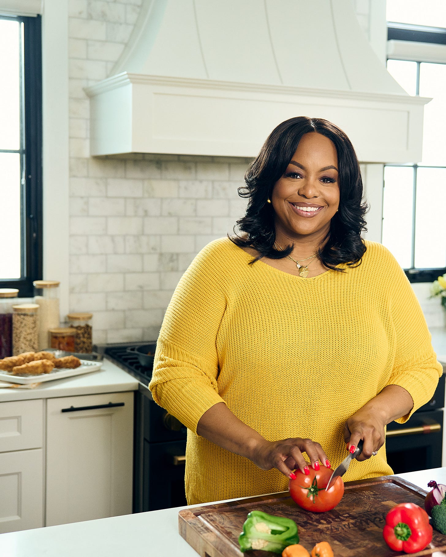 Kardea Brown in a white kitchen wearing a bright yellow sweater in the middle of slicing a tomato on a wooden cutting board