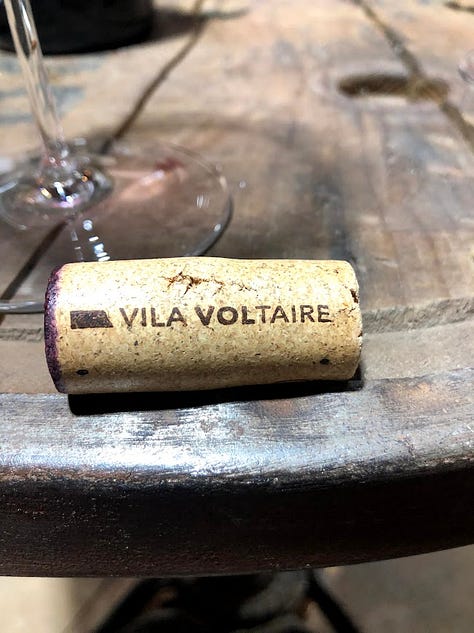 A gallery of cork closures: a natural cork branded 'Vila Voltaire' resting on a wine barrel table, a cork being pulled from a bottle with a waiter's corkscrew, and a cluster of Champagne corks and cages from various producers