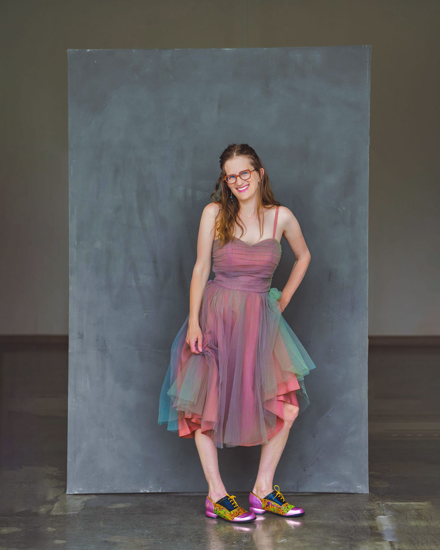 A white woman in a dusty-pink vintage gown with green tulle. She has glasses and cute shoes.