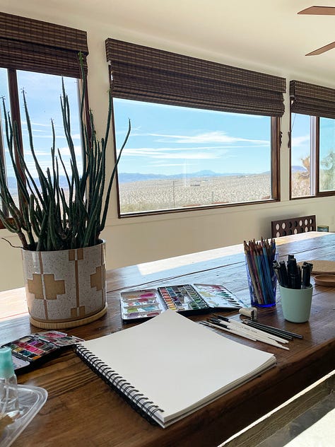 Art supplies on table with view of the desert, pinks and purples in the sky for sunset, a joshua tree at sunset, a pink house in the desert that matches the pink colors in the sky