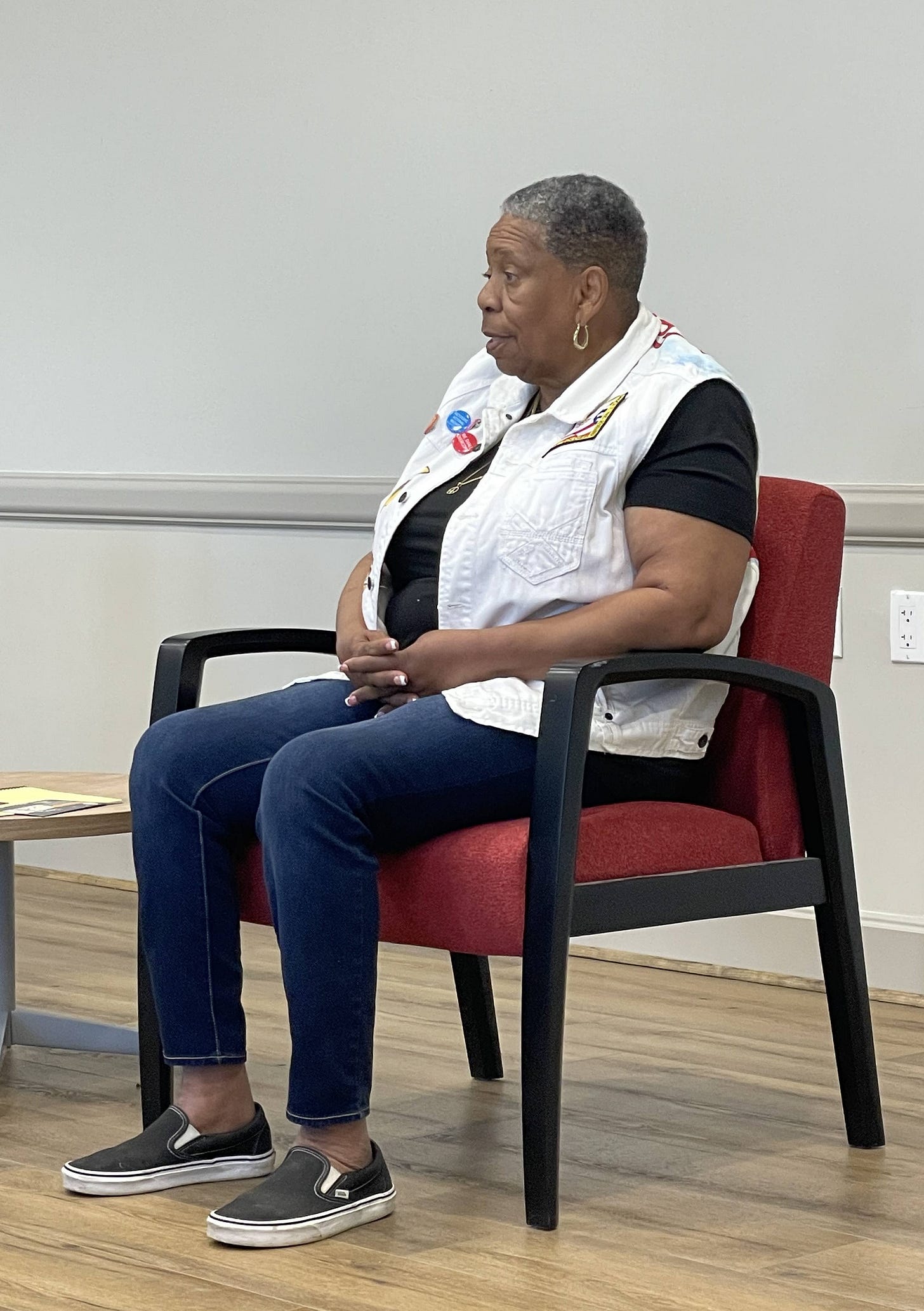 Black woman with close-cropped silver black hair wearing a white jeans vest, black t-shirt, and blue jeans, addresses a crowd while seated on a red chair.