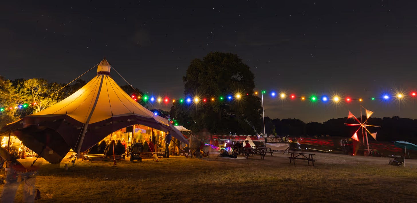 PIcture of some tents in a field in the night-time, with coloured string lights hanging around the area.
