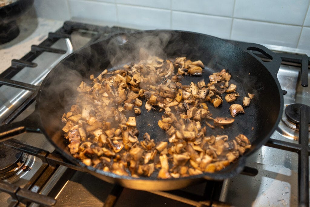 Steam rising from finely chopped mushrooms getting a hard, dark sear in a black cast iron skillet over a gas burner.