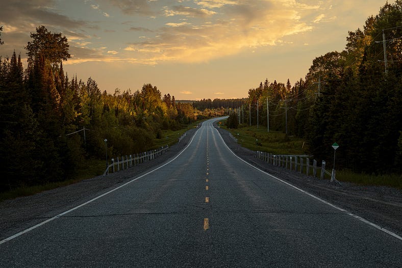 Tree lined paved road with yellow broken line down the middle. Road is empty and the sky is dusk