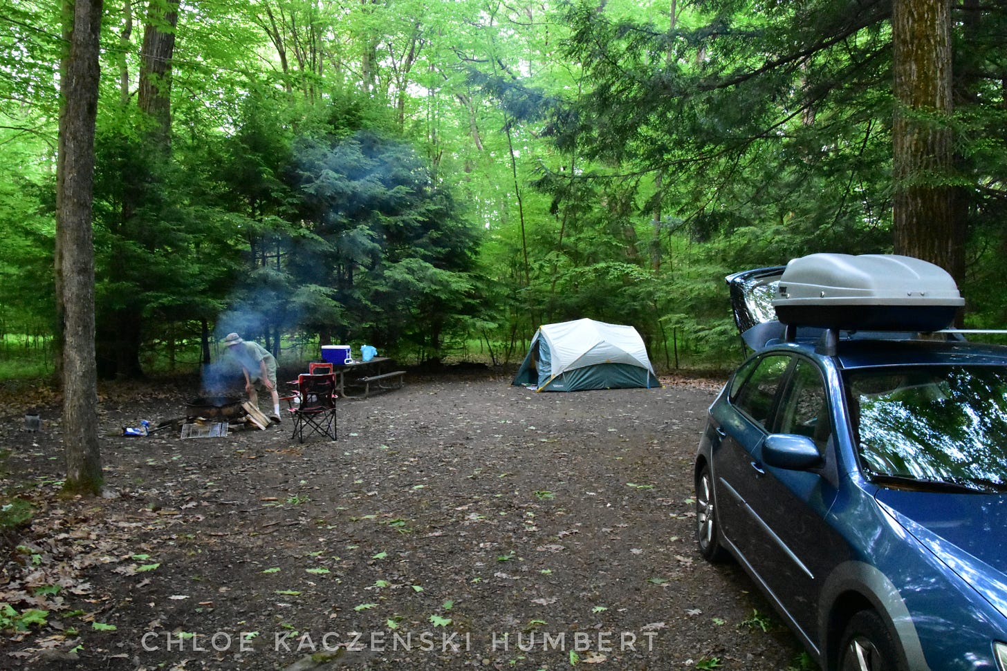 Camping in the Allegheny National Forest, Pennsylvania. June 2, 2018. Photo by Chloe Kaczenski Humbert. Photo shows a campsite surrounded by lush trees with a tent, someone building a fire in a fire pit, and a car with the hatch open and a cargo carrier on top.