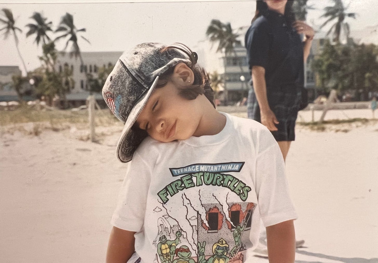 Photo of young girl standing on the sand with her mother and buildings and palm trees in the background.