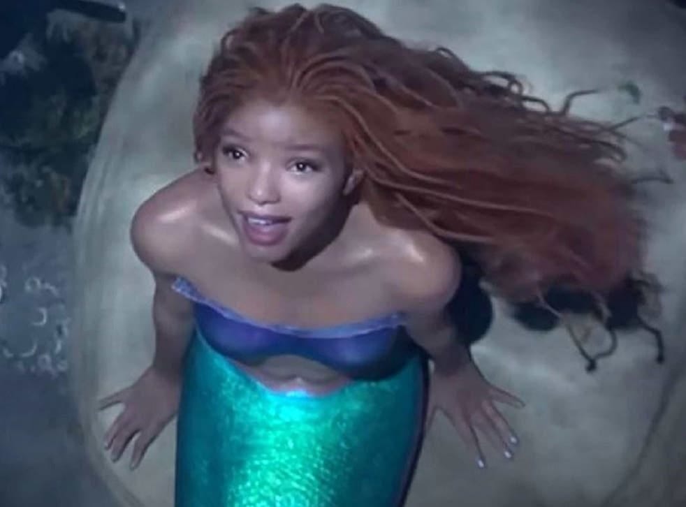 Young woman with brown skin and long, thick, dark hair, wearing a blue strapless top and whose bottom half is a green scaly fish fin, sitting on a large rock and looking up into the sky, singing