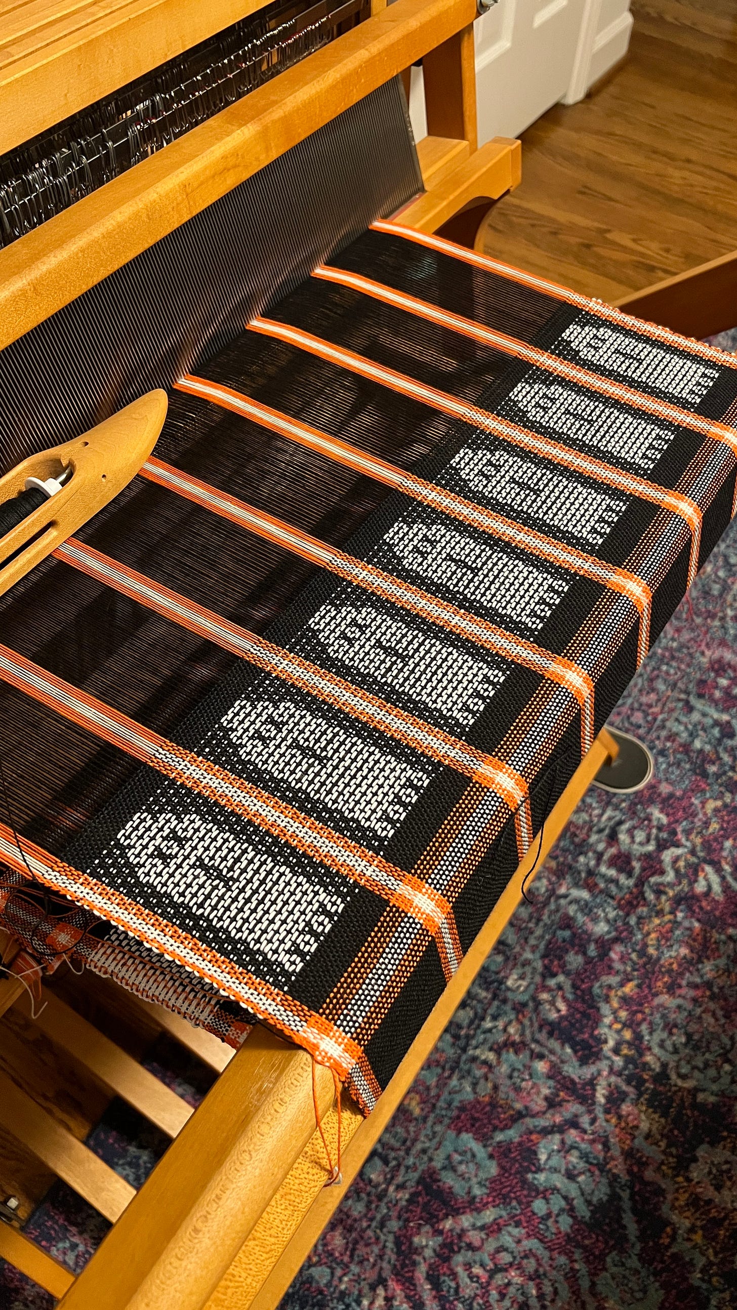 a photo of a floor loom with black and orange and white stripes with ghosts woven in