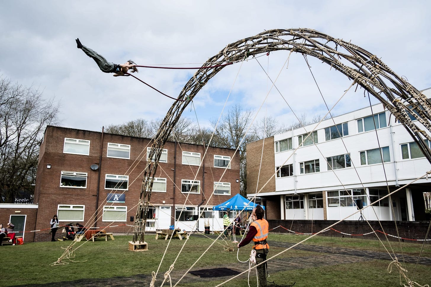 A performer swings high in the air suspended from an arch made from bamboo, with university buildings in the background
