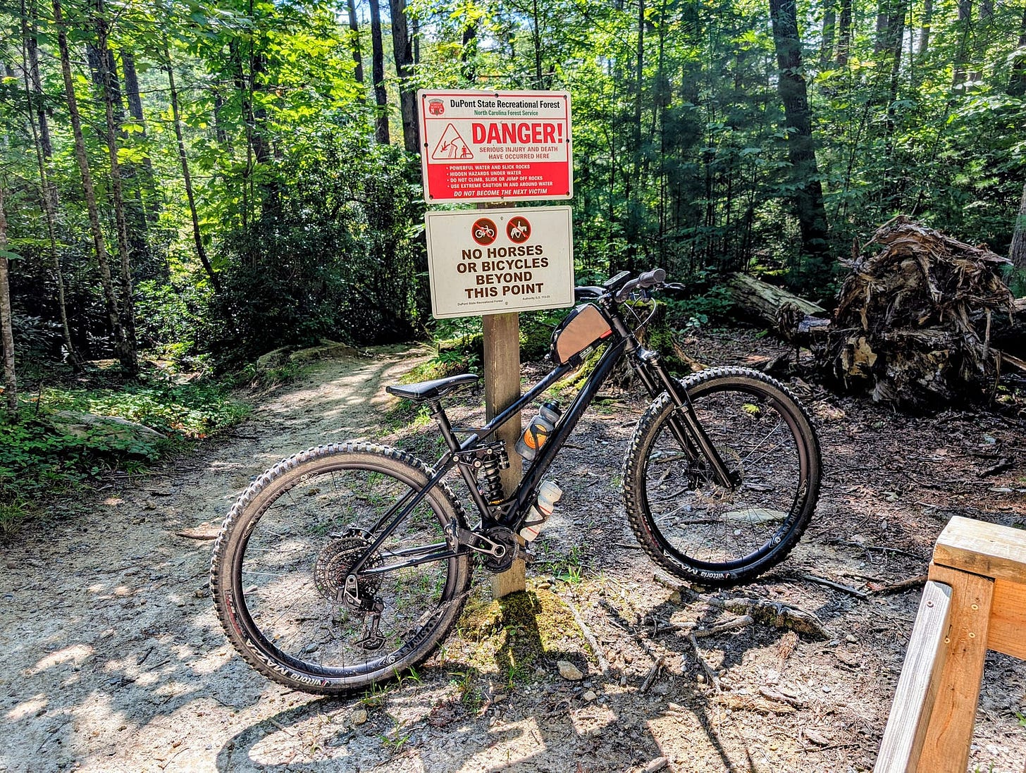 A matte-black REEB mountain bike leans against a trail signpost in DuPont State Recreational Forest. The sign warns of dangerous conditions ahead and prohibits horses or bicycles beyond that point. Sunlight filters through dense green forest, illuminating the sandy trail that continues on foot past the sign. The scene captures a pause before transition — from wheels to walking — in the heart of rugged terrain. A matte-black REEB mountain bike leans against a trail signpost in DuPont State Recreational Forest. The sign warns of dangerous conditions ahead and prohibits horses or bicycles beyond that point. Sunlight filters through dense green forest, illuminating the sandy trail that continues on foot past the sign. The scene captures a pause before transition — from wheels to walking — in the heart of rugged terrain.