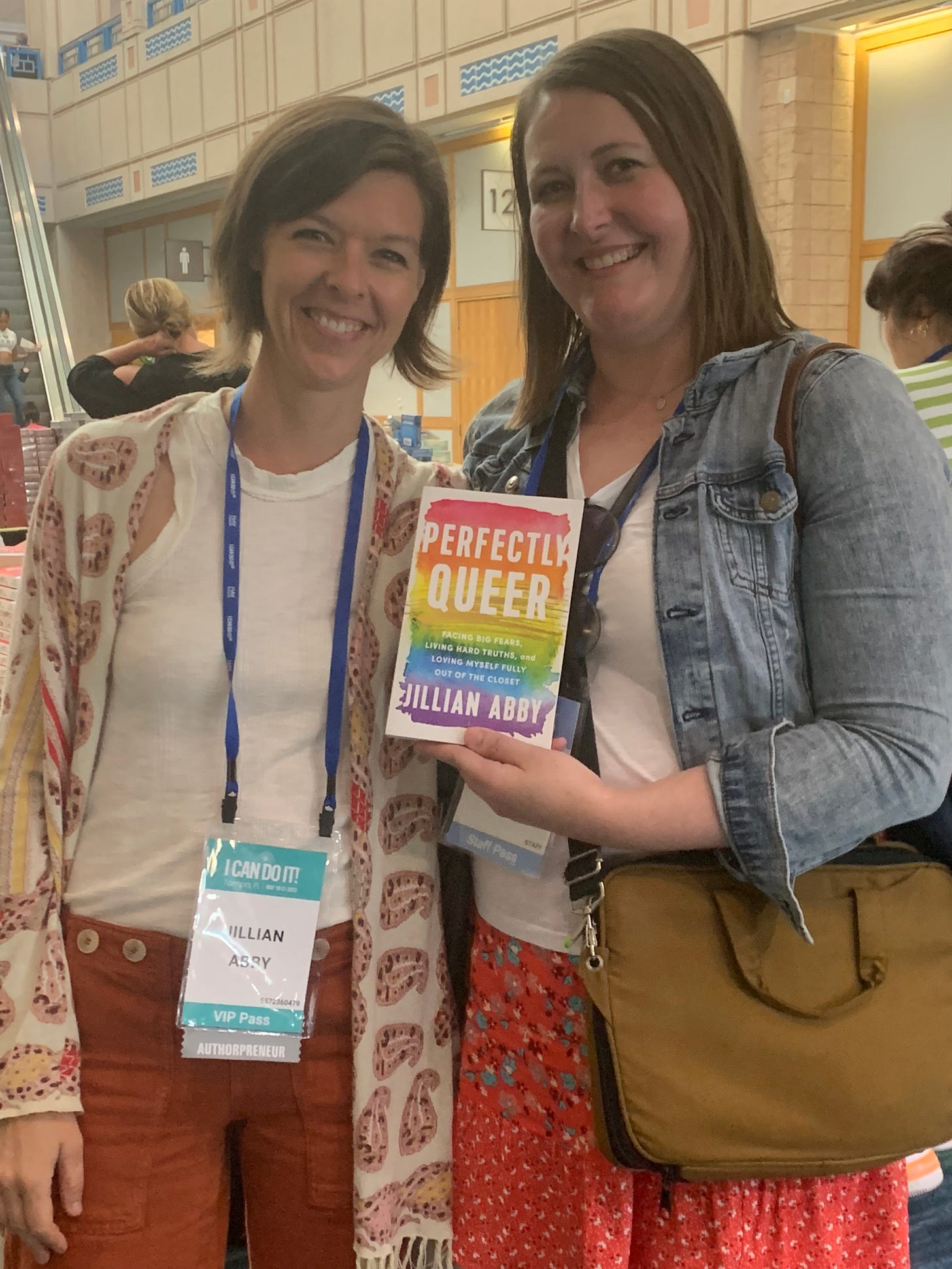 Two women, both with shoulder-length brown hair, smile together holding a copy of the book Perfectly Queer from Hay House publishing.
