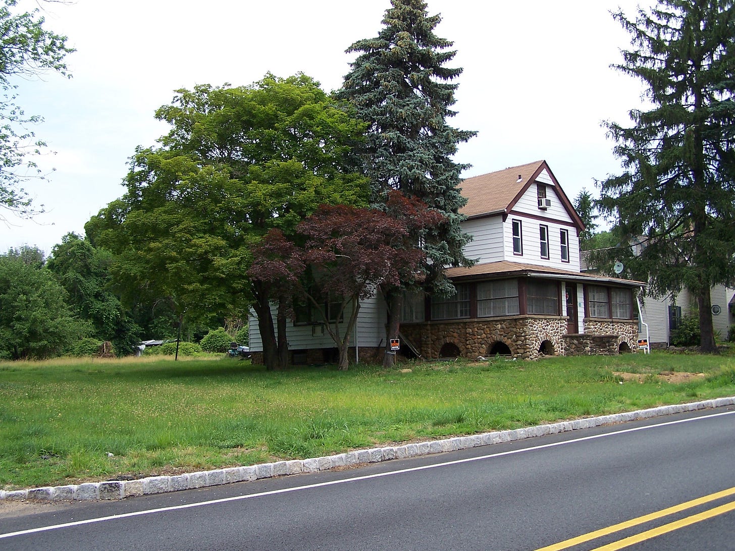 Photo from across the street of a very tired-looking farmhouse. Next to it there are three spectacular trees: a 40' tall green-leaf maple, a 20' red leaf maple, and a 60' blue spruce.