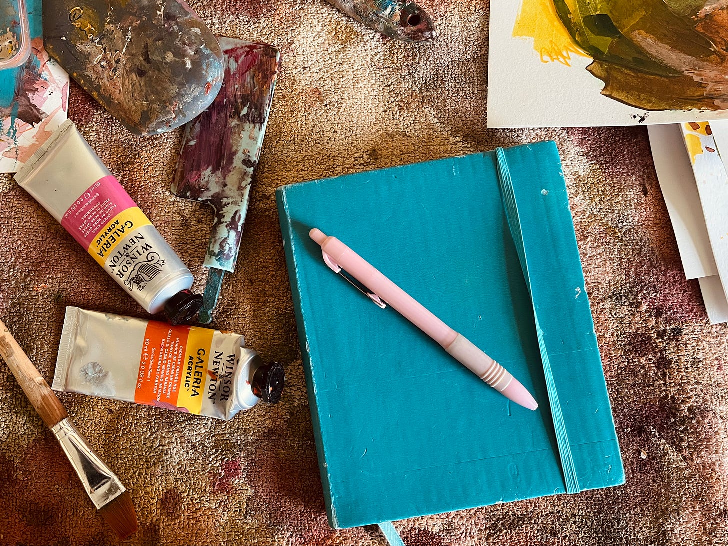A painter’s workspace filled with creativity—a well-used journal with a pink pen resting on top, surrounded by acrylic paint tubes, brushes, and palette knives. The textured surface beneath is covered in layers of dried paint, reflecting the messy and vibrant process of art-making.