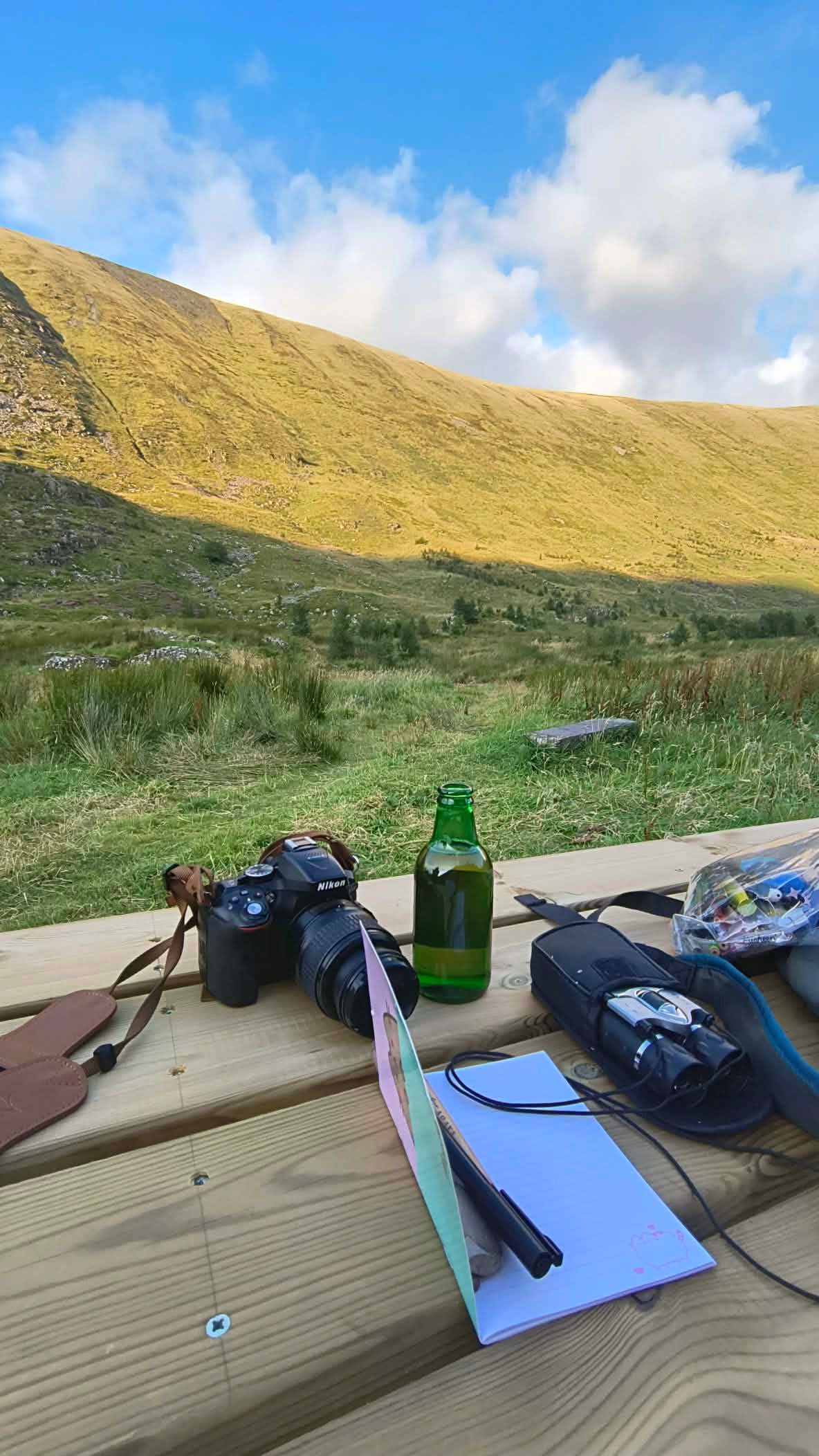 at the base of a glowing lowland hills is a bench, on it sits a camera, binaculars, notepad and small beer