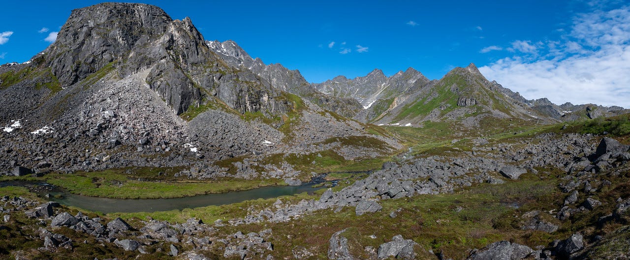 A panoramic view of a rocky alpine valley under a bright blue sky scattered with thin white cloud. At left, a dark rock buttress rises steeply above slopes of grey scree. A jagged line of pinnacled peaks runs across the middle distance, small snow patches held in the shaded couloirs, green vegetation climbing the lower slopes. The foreground is a green tundra valley floor strewn with grey boulders, a calm, green-tinted creek curving through the center of the frame. Full summer light, most of the snow gone. A panoramic view of a rocky alpine valley under a bright blue sky scattered with thin white cloud. At left, a dark rock buttress rises steeply above slopes of grey scree. A jagged line of pinnacled peaks runs across the middle distance, small snow patches held in the shaded couloirs, green vegetation climbing the lower slopes. The foreground is a green tundra valley floor strewn with grey boulders, a calm, green-tinted creek curving through the center of the frame. Full summer light, most of the snow gone.