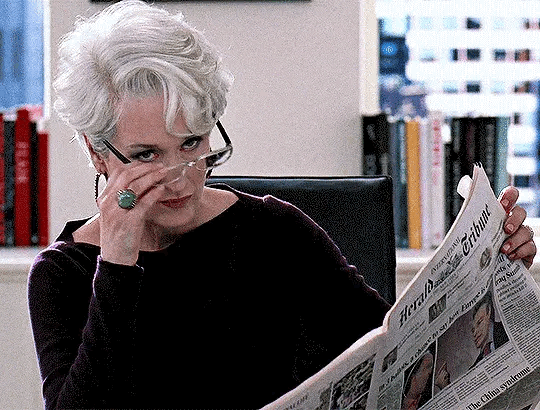 This may contain: a woman sitting in an office chair reading a newspaper while holding her hand to her face