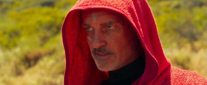 Julian McMahon in Red Hood Alt text: Close-up of Julian McMahon in “The Surfer” (2024), wearing a red hooded towel with a stern, intense expression under bright coastal light.