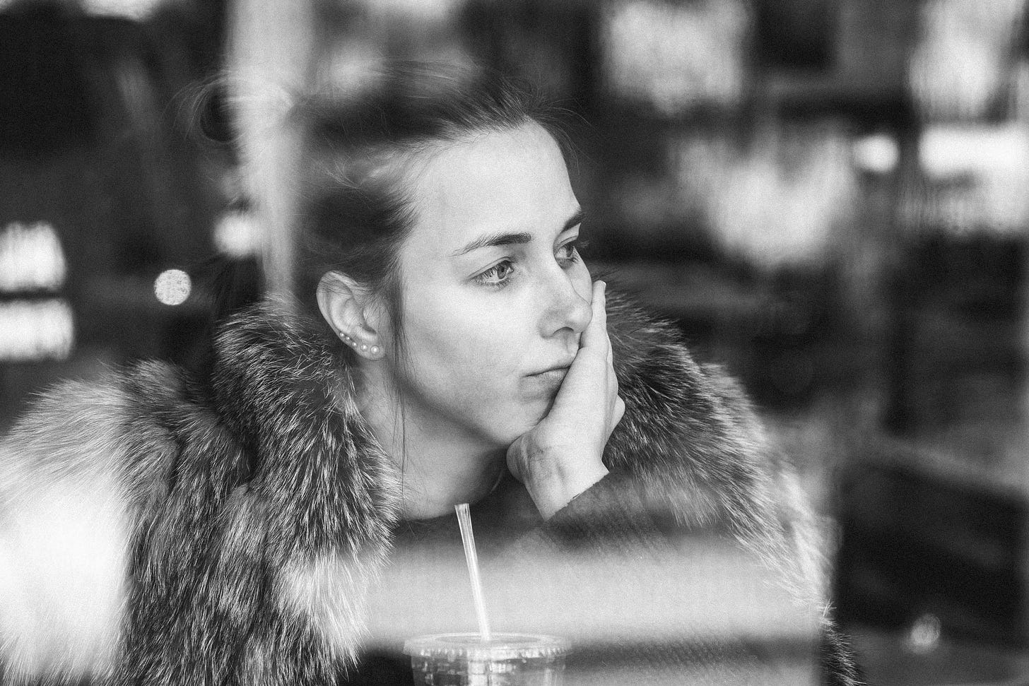 A black and white photo of artist CJ Hendry seen through a window. She is wearing a fur coat and rests her head on her hand with a pensive expression. The glass has prominent light reflections, creating a layered and contemplative effect over the image.