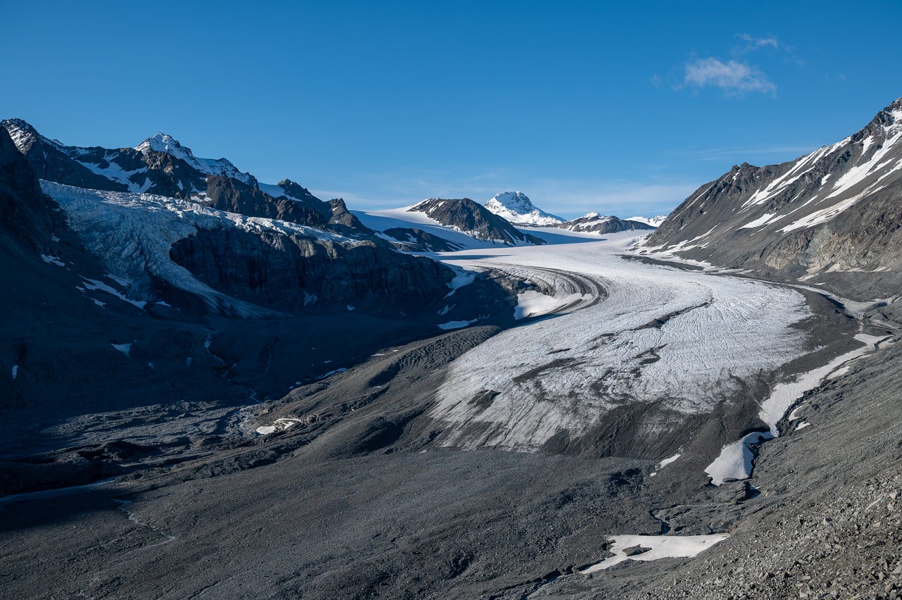 From atop a dark lateral moraine, a long tongue of grey-white glacier ice curves down its valley, streaked with dark medial bands of debris, flanked by rock walls and rising to snow-covered peaks and a rounded snowy dome at its head, under a clear deep blue sky with a single wisp of cloud.