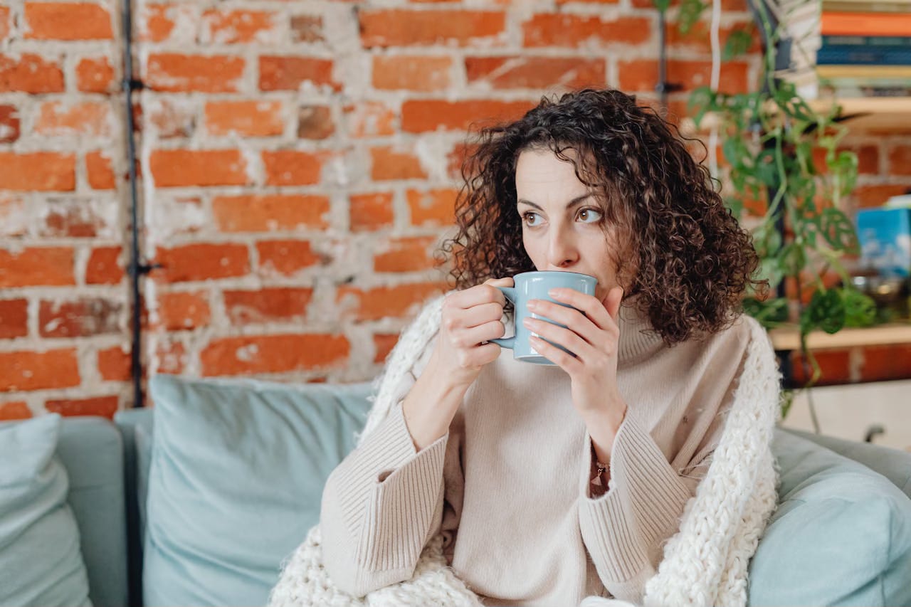A woman sits on a light aqua sofa next to a brick wall sipping coffee or tea from a aqua cup.