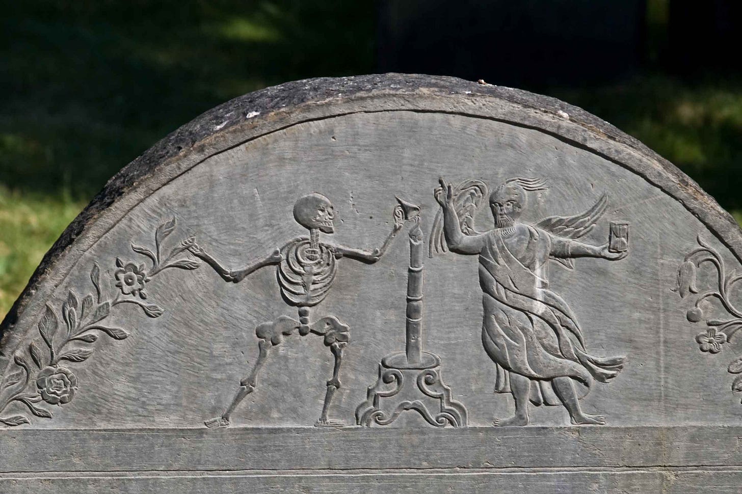 Top of a gravestone showing carved skeleton and human figure interacting