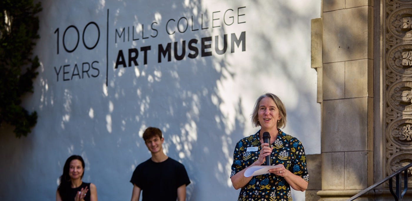 Mills College Art Museum Director Stephanie Hanor speaking into a microphone in front of wall displaying the text 'Mills College Art Museum: 100 Years.' Mills College Art Museum Director Stephanie Hanor speaking into a microphone in front of wall displaying the text 'Mills College Art Museum: 100 Years.'