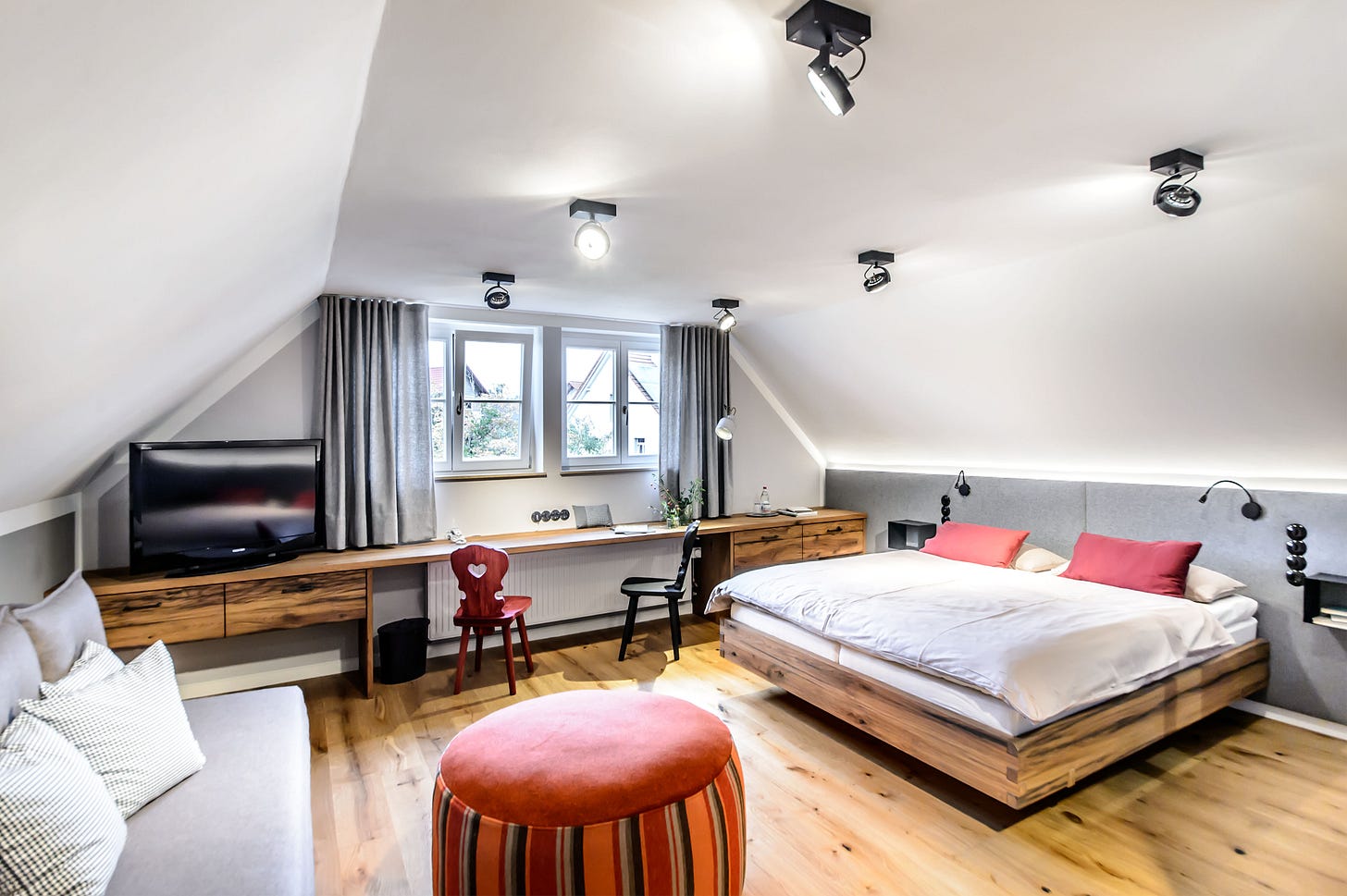 A large hotel room with slanted ceilings and furnishings in natural wood, and red and grey fabrics.