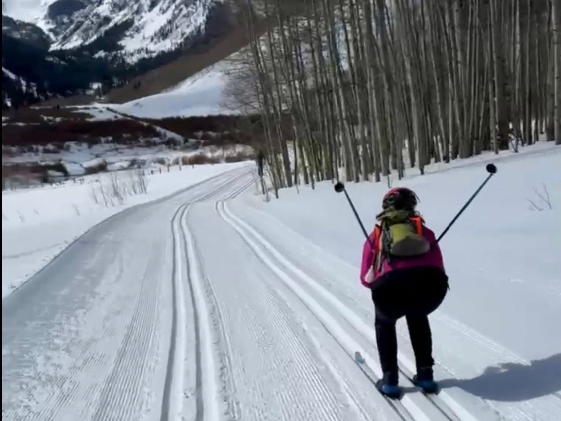 A skier in a bright pink jacket and black pants glides down a freshly groomed Nordic trail, poles raised behind them in a V shape. Snow-covered mountains rise in the background, framed by a stand of tall, leafless aspen trees on the right. The scene is peaceful and expansive, with blue sky peeking through the peaks.