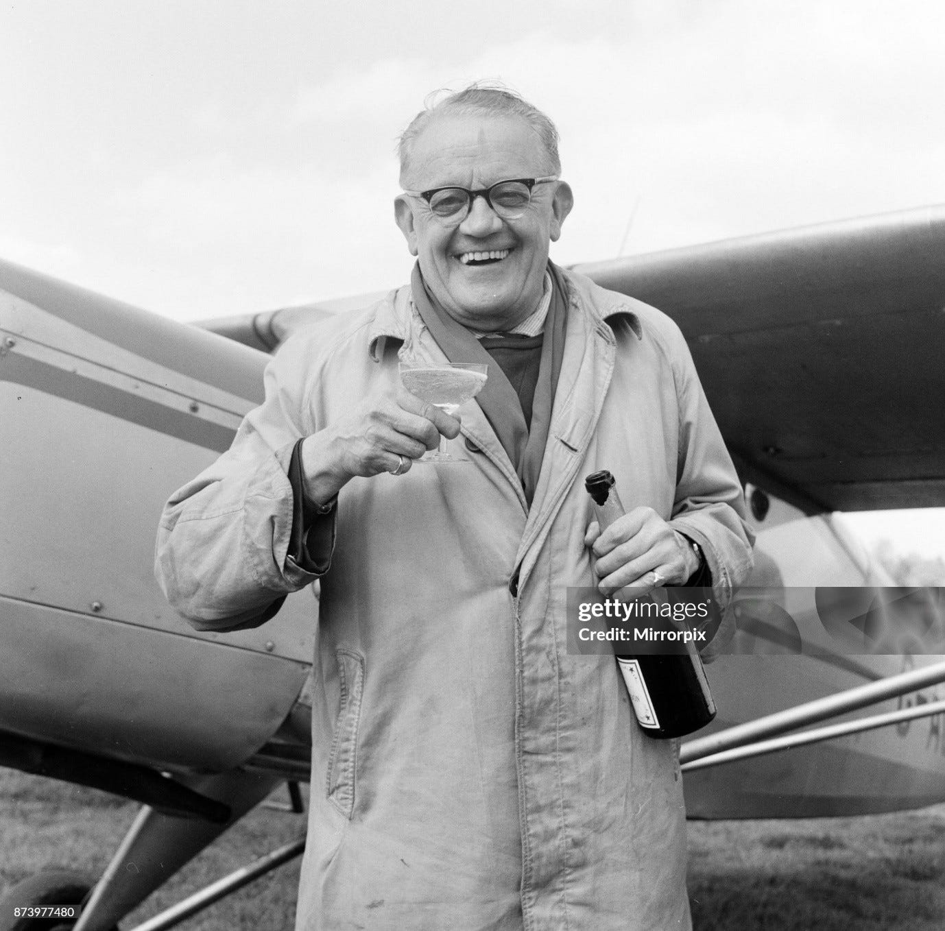 b/w photo of a man in a 1950s raincoat standing by a plane with a full champagne glass in one hand, bottle in the other, grinning proudly at camera. And rightly so