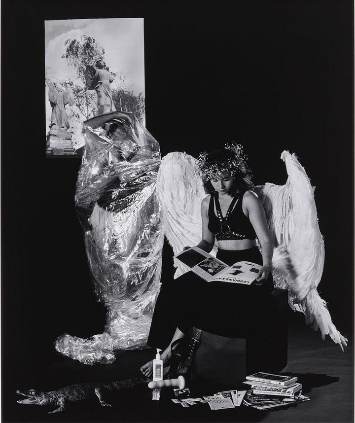 1) A black-and-white photo of a barefoot person in large white angel wings and a floral crown, sitting on a box and reading a magazine. On the wall behind them, there is a black and white photograph of angelic statues. In front of the portrait, there is another angel wrapped up in clingwrap. There is a faux alligator on the floor, surrounded by safe sex magazines and pamphlets. There is also a dildo and a bottle of lotion on the floor, in front of the angel. The scene feels surreal. 2) Two people in elaborate costumes face each other. One is sitting wearing large, white angel wings, while the other stands. A statue photo is visible behind the person who just emerged from the clingwrap. The clingwrap sits around their feet on the ground, next to the faux alligator. The person who was wrapped up is wearing a leather chest harness, a long frilly shirt covering their crotch, sunglasses, and latex gloves on both hands. They also have dark angel wings, with pieces of metal hanging off of them in several places. The person sitting down is looking over their wings to look at the other angel. The floor in front of them is still full of magazines, pamphlets, books, and a dildo with some lotion/lube. This photo is in black and white.
