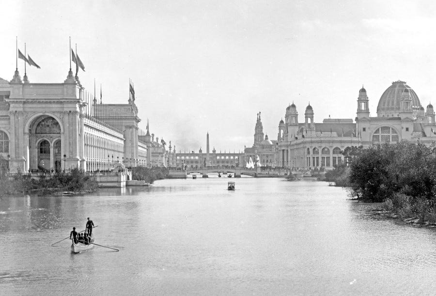 Gondoliers carry passengers across a lagoon, with several large buildings in the background.