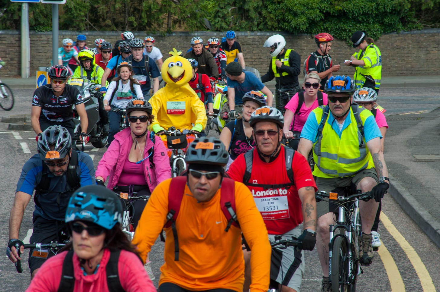 A colorful group of cyclists are driving torwards the camera. While most people are wearing standard cycling attire, one person is masked as Big Bird of Sesame Street.