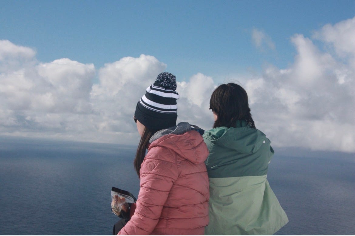 A landscape view of two people in winter jackets looking out over a blue sky with white clouds