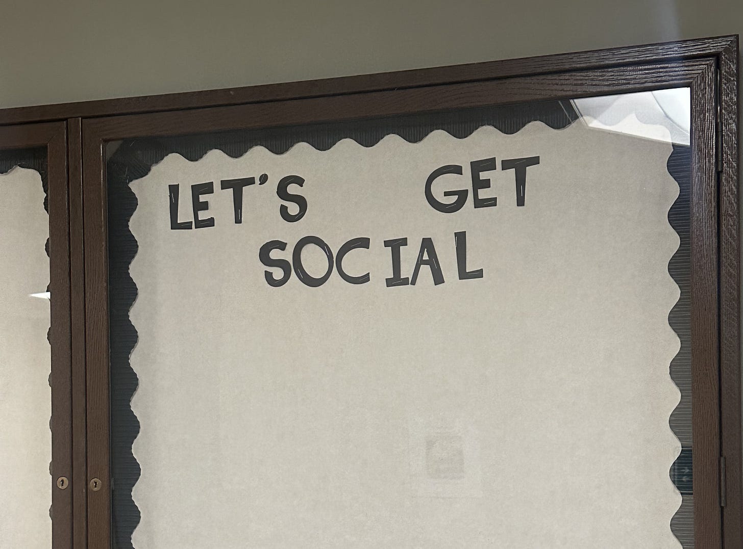 Bulletin board with scalloped edge paper and black text reading "LET'S GET SOCIAL" in a wooden frame, set against a plain wall.