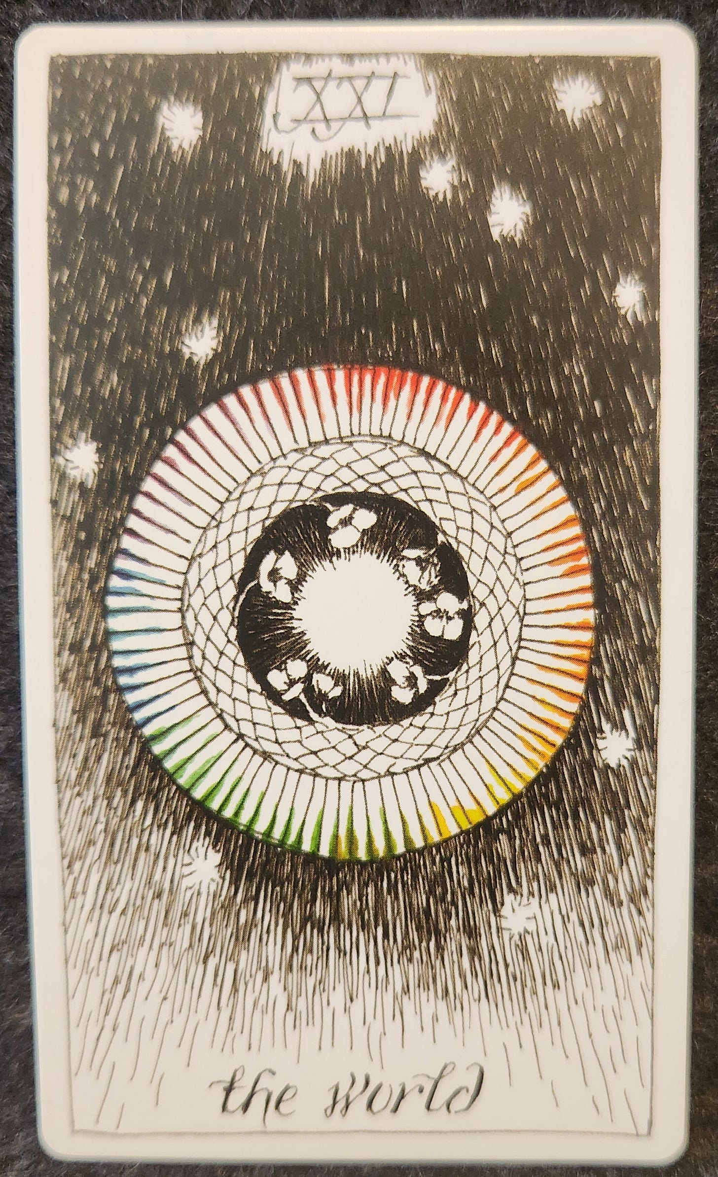 A tarot card labelled XXI the world. Nested circles, white, surrounded by a black circle of white flowers, surrounded by a patterned black and white circle, surrounded by a wider circle outlined in colours of the rainbow. The background is black with stars. 