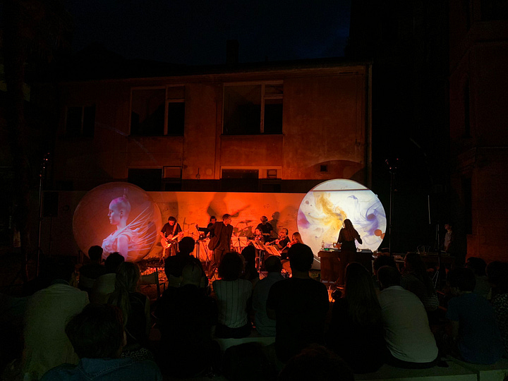 Nell'immagine si vede il gruppo musicale di A. Zannier suonare di sera davanti al pubblico, con una scenografia di luci e installazioni