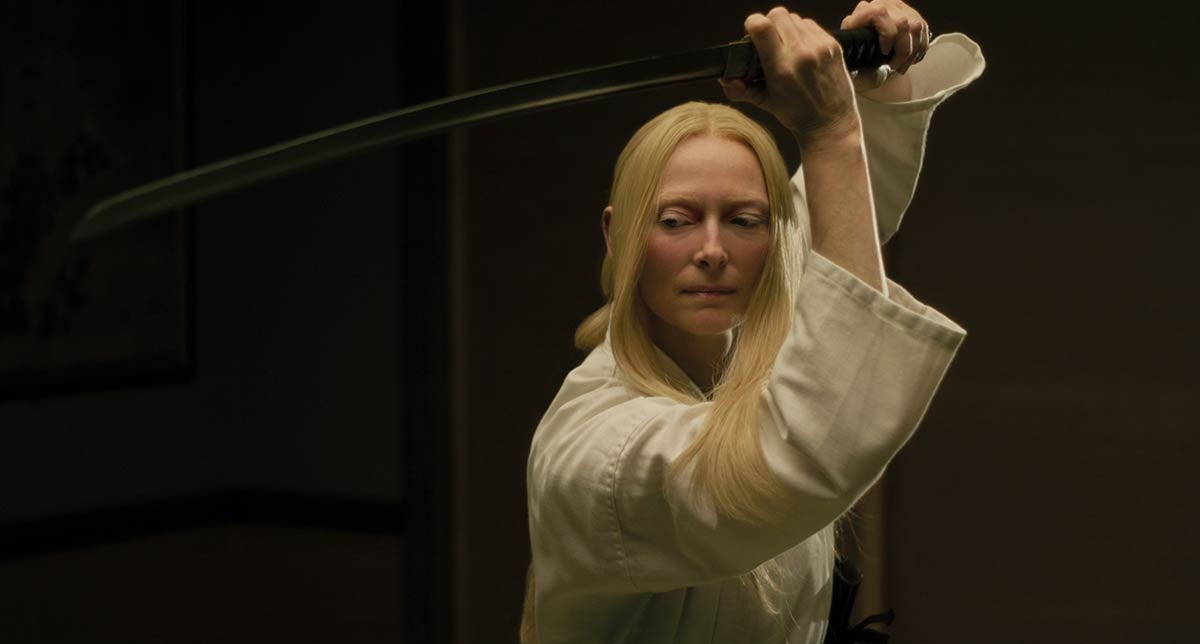 Tilda Swinton in white martial arts clothes, hair down, wielding samurai sword above her head