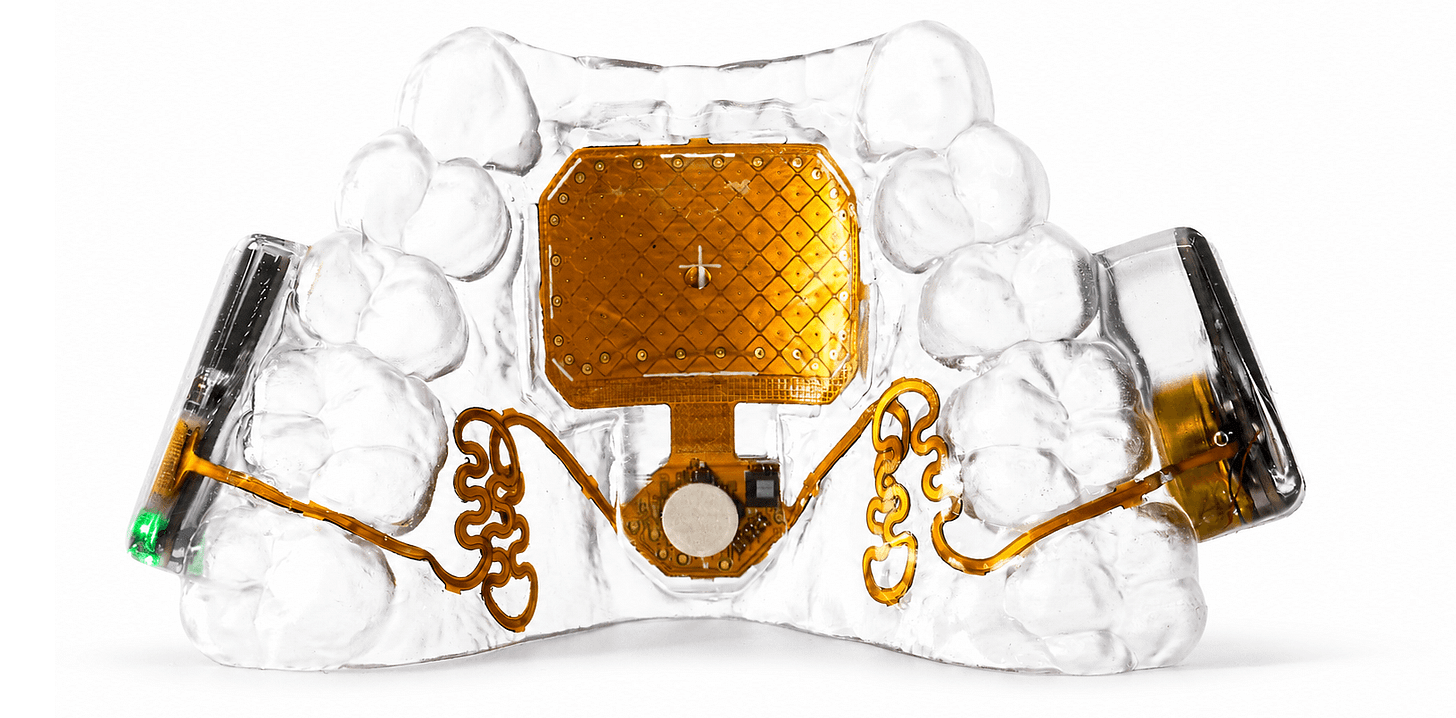 The MouthPad seen from the front: a clear custom-fit retainer with a gold touchpad at its center and gold flexible circuitry curving out to the side electronics, one of which shows a small green status light, lit against a white field.