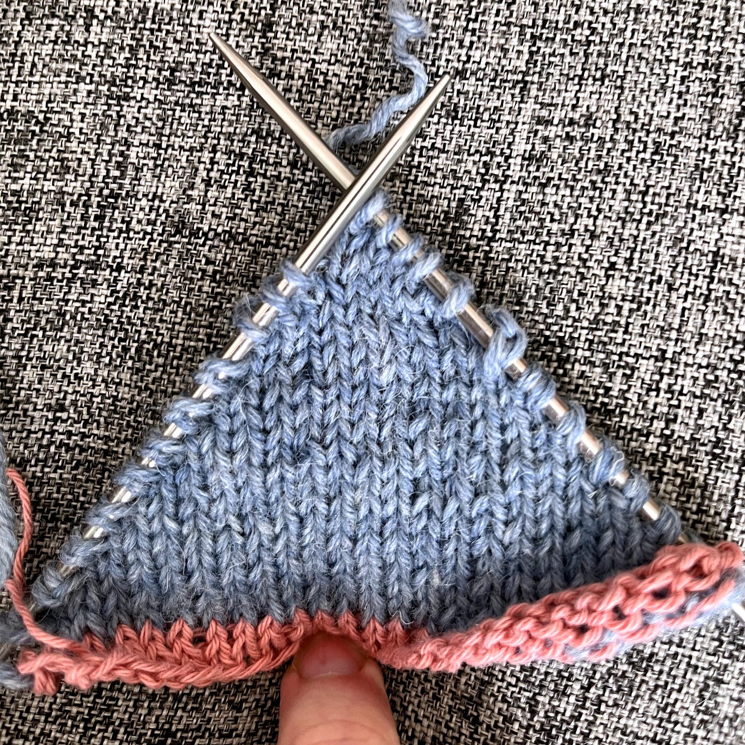 A triangular swatch of grey stocking-stitch knitting on double-pointed needles, with a pink crochet provisional cast-on visible along the base. A triangular swatch of grey stocking-stitch knitting on double-pointed needles, with a pink crochet provisional cast-on visible along the base.