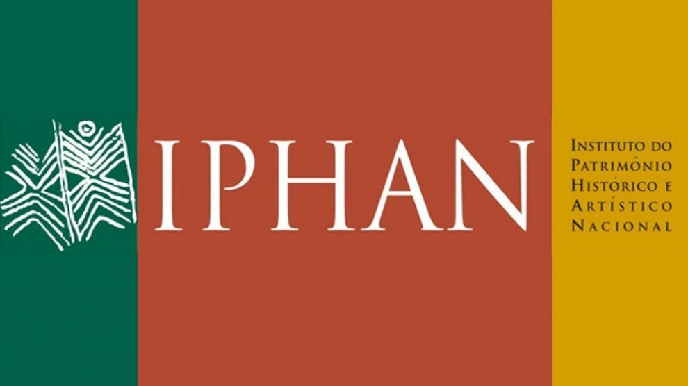 descrevemospravocê  Imagem com o logotipo do IPHAN – Instituto do Patrimônio Histórico e Artístico Nacional, distribuída em três faixas verticais coloridas.  Na faixa da esquerda, com fundo verde escuro, há um símbolo branco que representa um grafismo indígena estilizado, remetendo a formas ancestrais e à arte rupestre brasileira. O desenho tem linhas diagonais e curvas que formam figuras simétricas, representando elementos do patrimônio cultural.  Ao centro, sobre um fundo em tom terroso avermelhado, está a sigla IPHAN, em letras brancas e serifadas, com espaçamento amplo e elegante.  Na faixa da direita, com fundo amarelo-ouro, está escrito em letras pretas, com destaque para as iniciais em maiúsculas: INSTITUTO DO PATRIMÔNIO HISTÓRICO E ARTÍSTICO NACIONAL.  As cores, a tipografia clássica e os elementos gráficos reforçam a identidade visual institucional do IPHAN, que está vinculado à preservação do patrimônio histórico e cultural brasileiro.