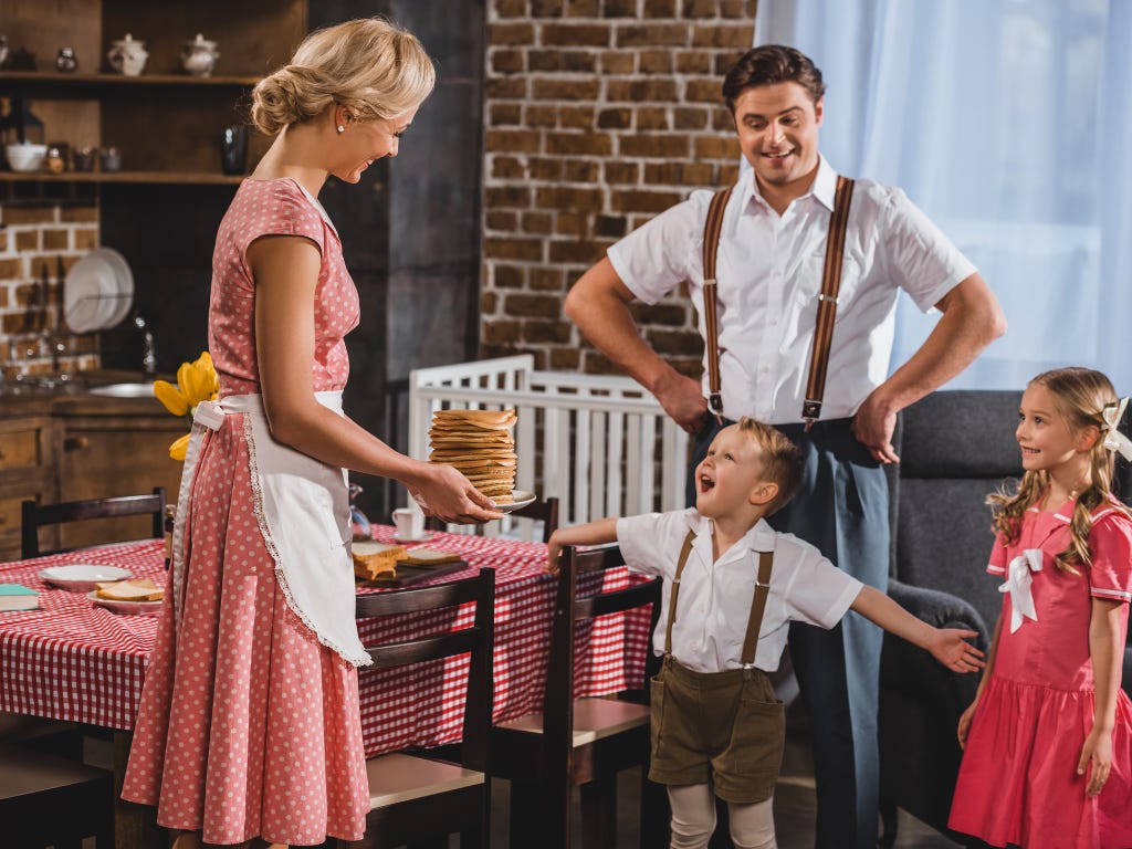 Gestileerde jaren-vijftigscène waarin een moeder in schort eten serveert aan twee kinderen terwijl de vader ernaast staat. Gestileerde jaren-vijftigscène waarin een moeder in schort eten serveert aan twee kinderen terwijl de vader ernaast staat.
