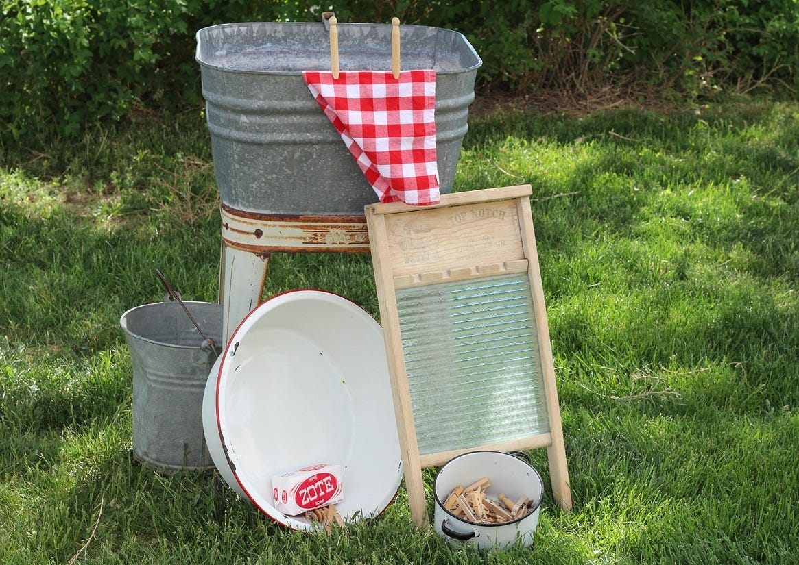 Washtub with towel hanging over the side, bucket, washboard and tin of clothes peg, all gathered on grass