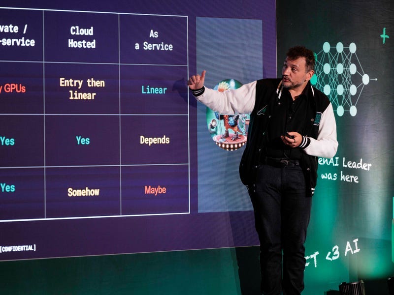 A person is standing and gesturing in front of a screen displaying a comparison table with columns labeled \ A person is standing and gesturing in front of a screen displaying a comparison table with columns labeled \