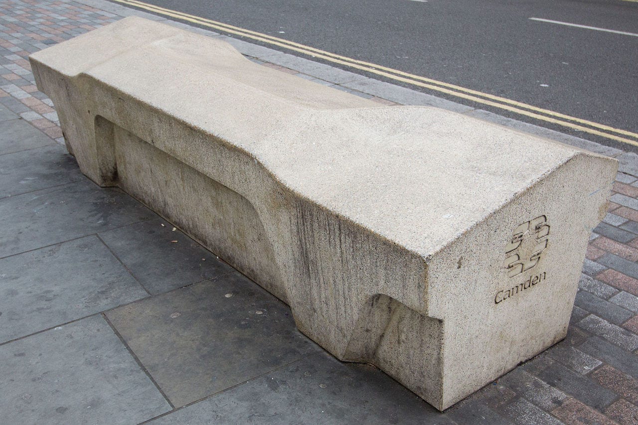 The "Camden Bench", one of the most notorious examples of civic hostile architecture masquerading as street furniture. Made of solid concrete, it is angled so that it is uncomfortable enough to deter anyone to sit on it for long periods of time; the multi-angled planes of the bench’s seating and absence of a back rest means you can not lie down or relax long enough to doze off; the solid concrete form factor prevents bags from being stored underneath for safekeeping. The message is "Sit down, but don't even think about relaxing or getting comfortable"