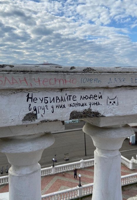 A weathered stone balustrade with white columns under a partly cloudy sky. Hand-painted graffiti in red and black reads "САНИ, техника любви, САНЯ СО" and "Не убивайте людей за чужие комиксы." A cat face emoji is drawn next to the text. The view shows a paved area with people walking below.