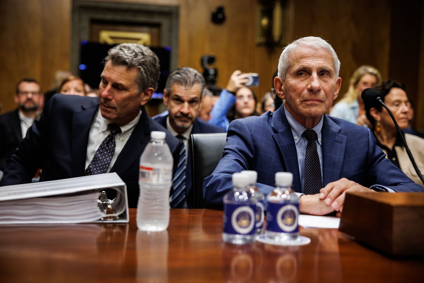 Anthony Fauci seated at the hearing table beside his legal team, with a thick binder and water bottles. Anthony Fauci seated at the hearing table beside his legal team, with a thick binder and water bottles.