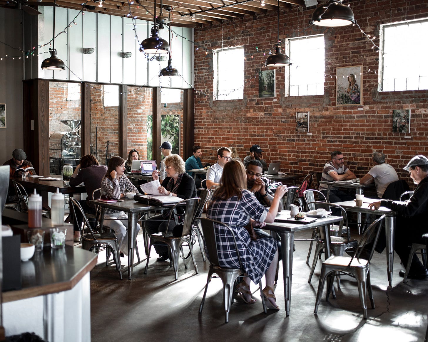 A busy coffee shop interior. Photographed from a corner looking out at customers sitting at table chatting, working on computers or reading. The far wall to the right is old brick, and to the left are framed floor to ceiling windows. Christmas lights and hanging lamps hang down from an uninsulated open ceiling.