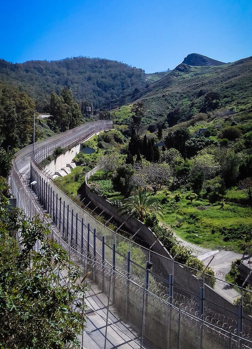 The Ceuta–Morocco border fence curves through a steep, green mountain valley. The Ceuta–Morocco border fence curves through a steep, green mountain valley.